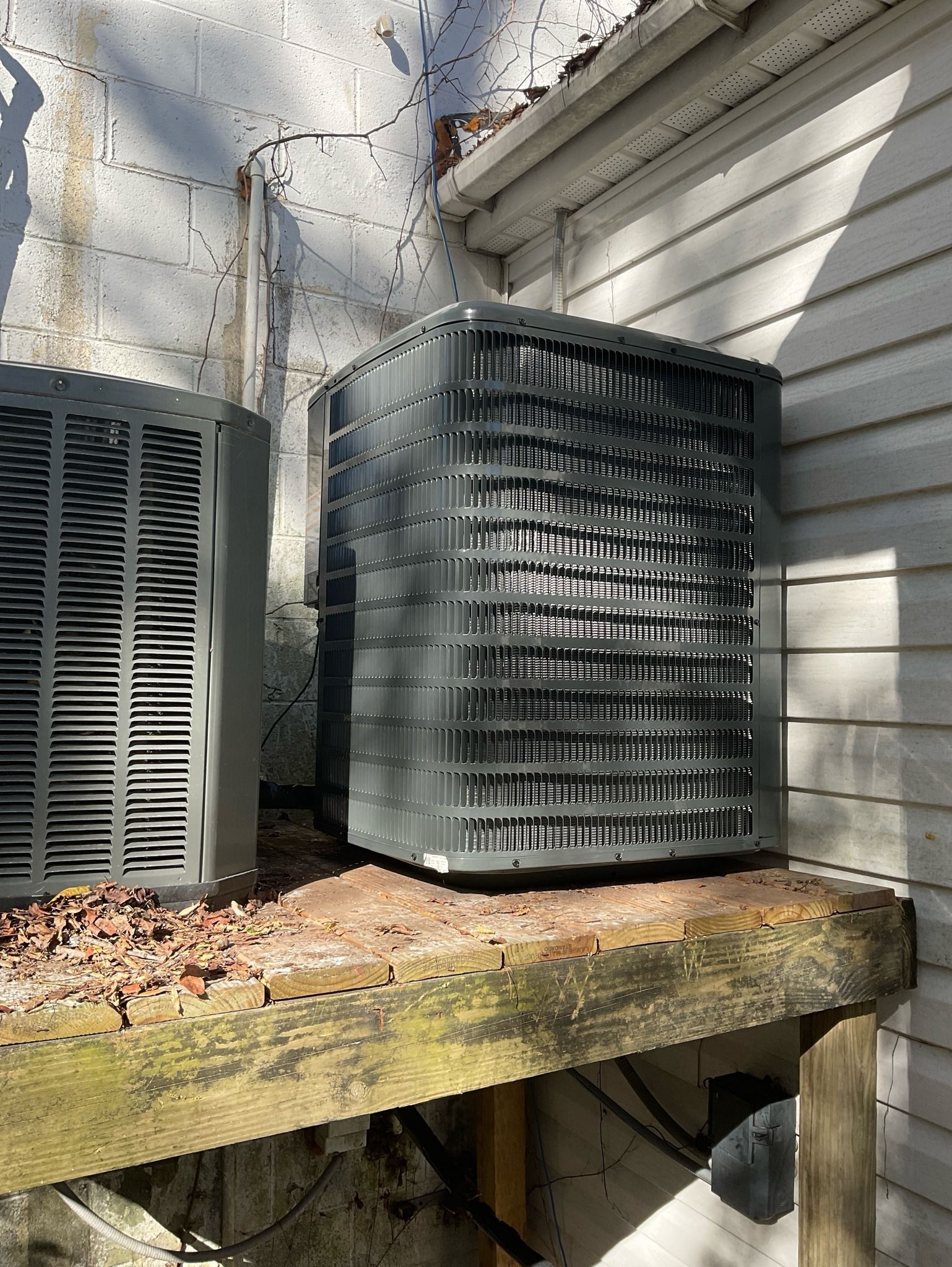Two air conditioners are sitting on a wooden shelf outside of a house.