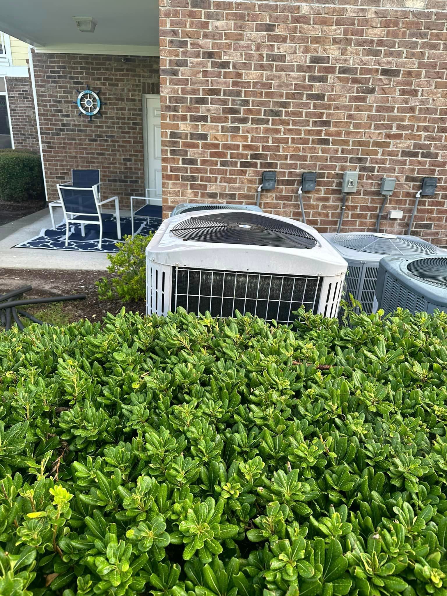 An air conditioner is sitting in front of a brick building next to a bush