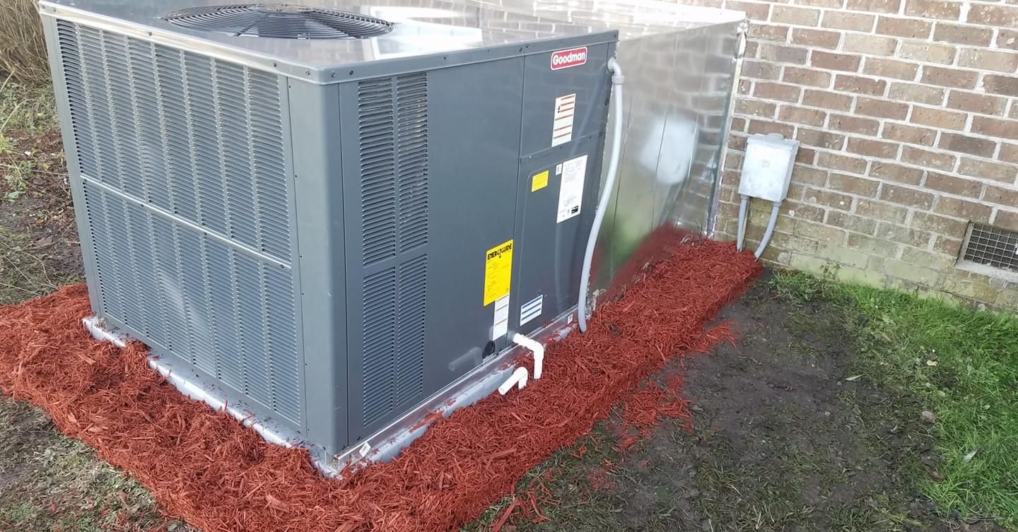 A large air conditioner is sitting on top of a pile of mulch next to a brick building