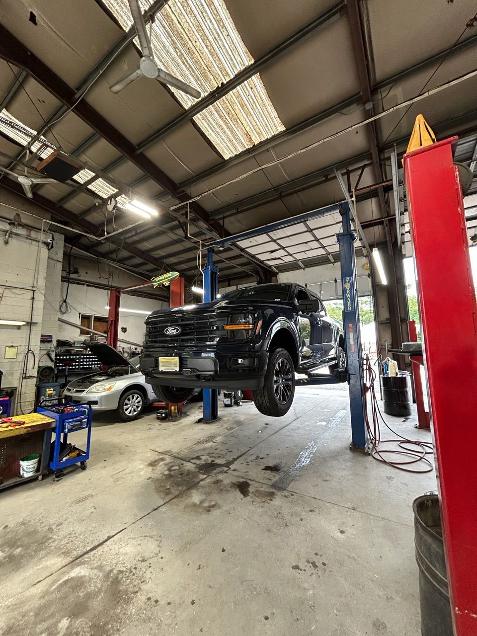 Black pickup truck lifted in a garage workshop, with another car and red lift columns nearby.