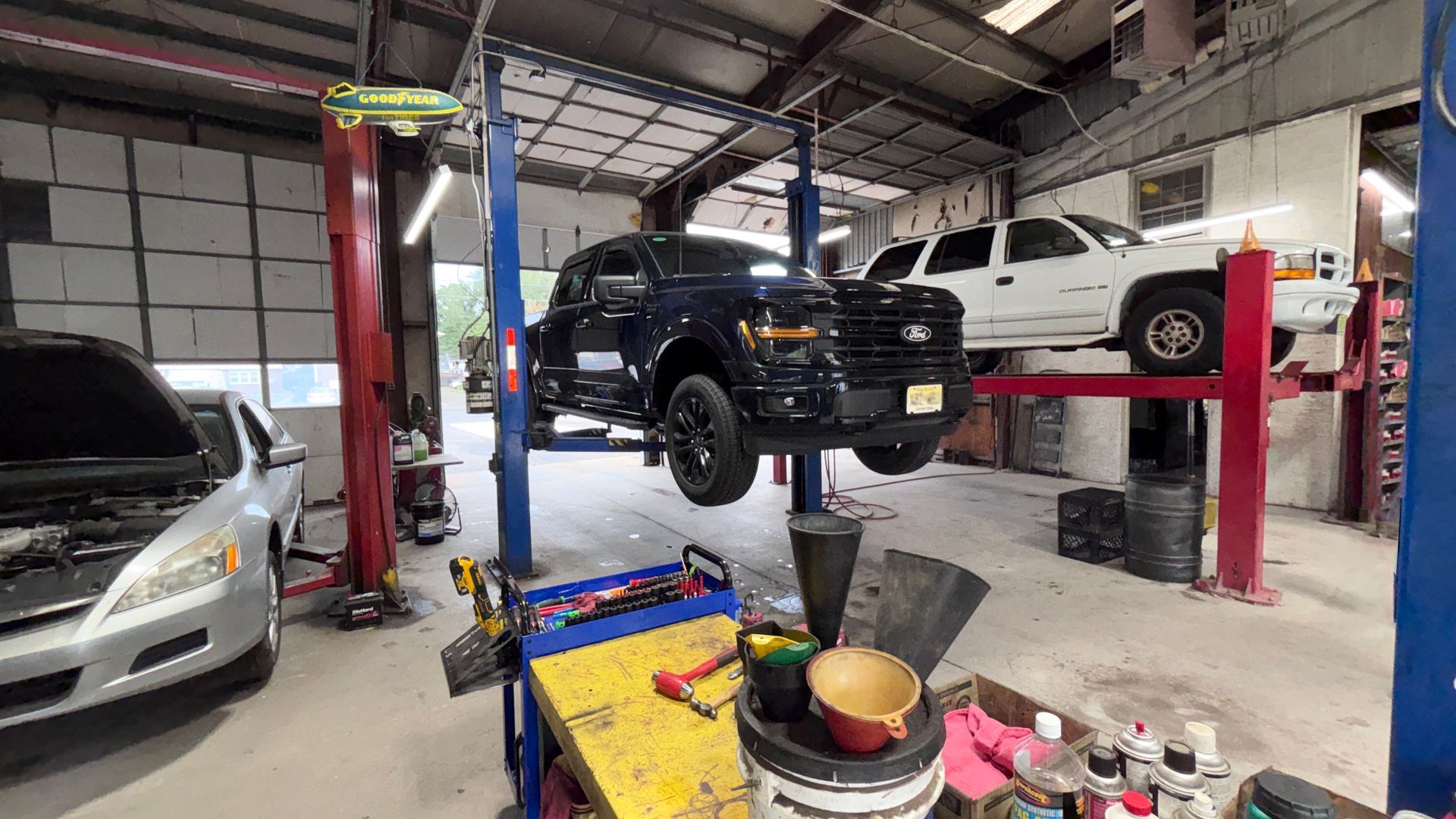 Auto repair shop with a black SUV on a lift, a white car beside it, and tools in the foreground.