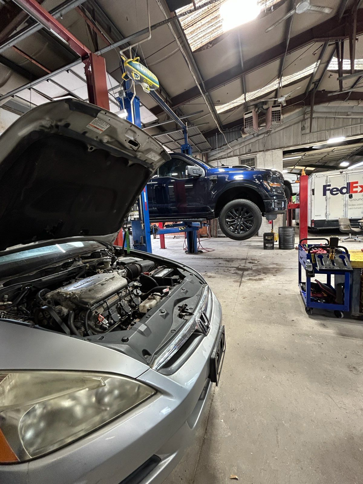 Auto repair shop with a silver car hood open and a blue pickup lifted on a hoist, mechanic working overhead