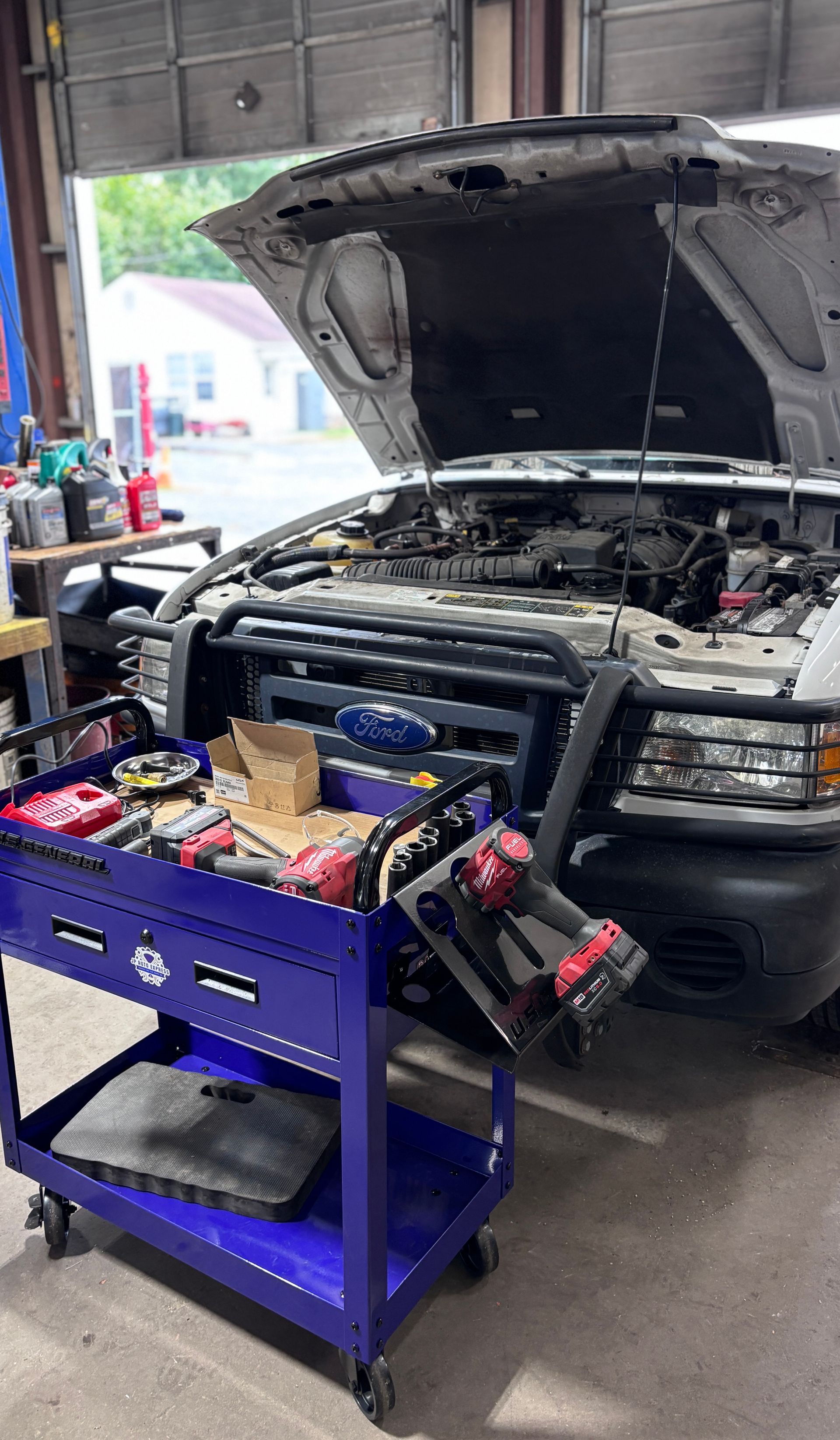 SUV in a garage with hood open, battery tester cart in front, and engine bay exposed for repair
