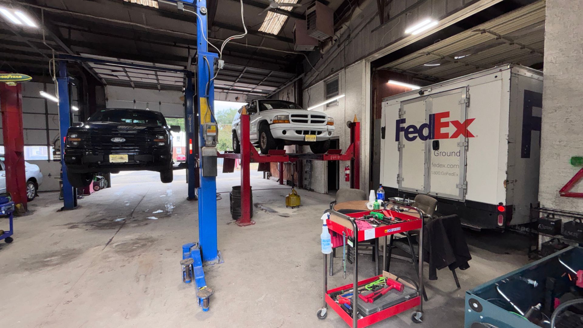 Auto repair garage with a lifted black truck, red car lift, and FedEx box truck inside.