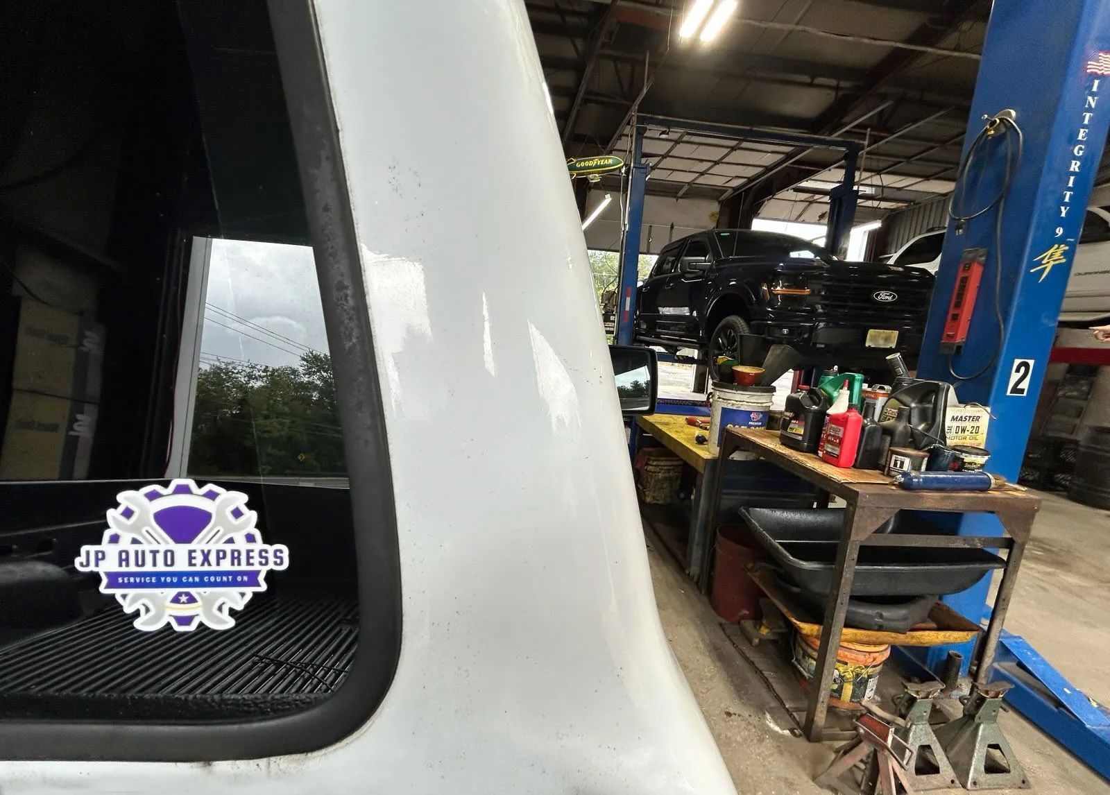 White vehicle in a garage with a car lift, tools, and a black SUV under repair.