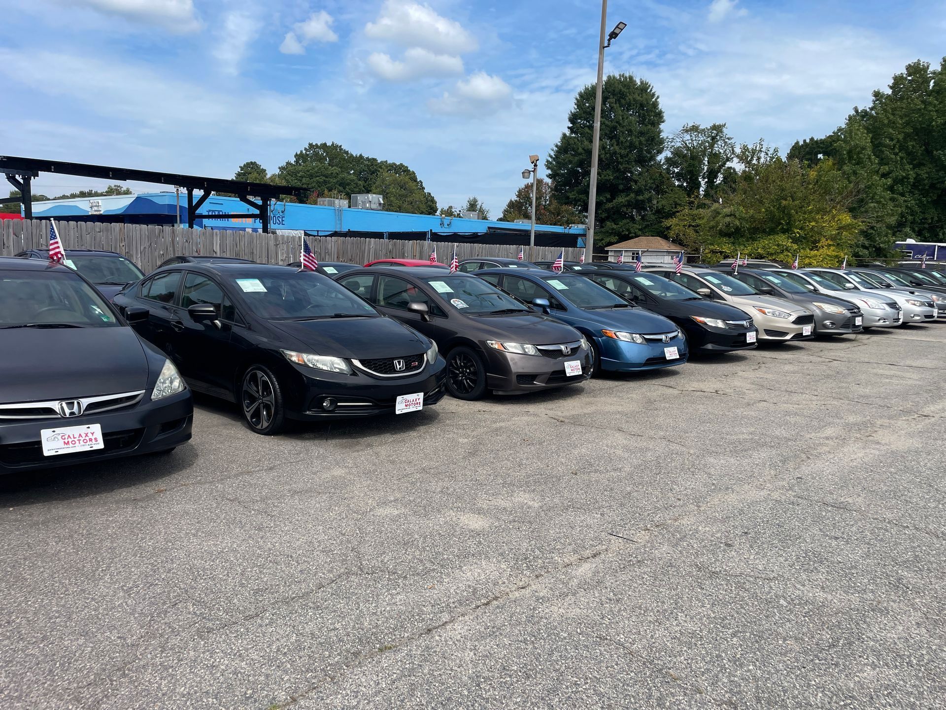 Cars parked in a row at a dealership on a gravel lot under a blue sky.