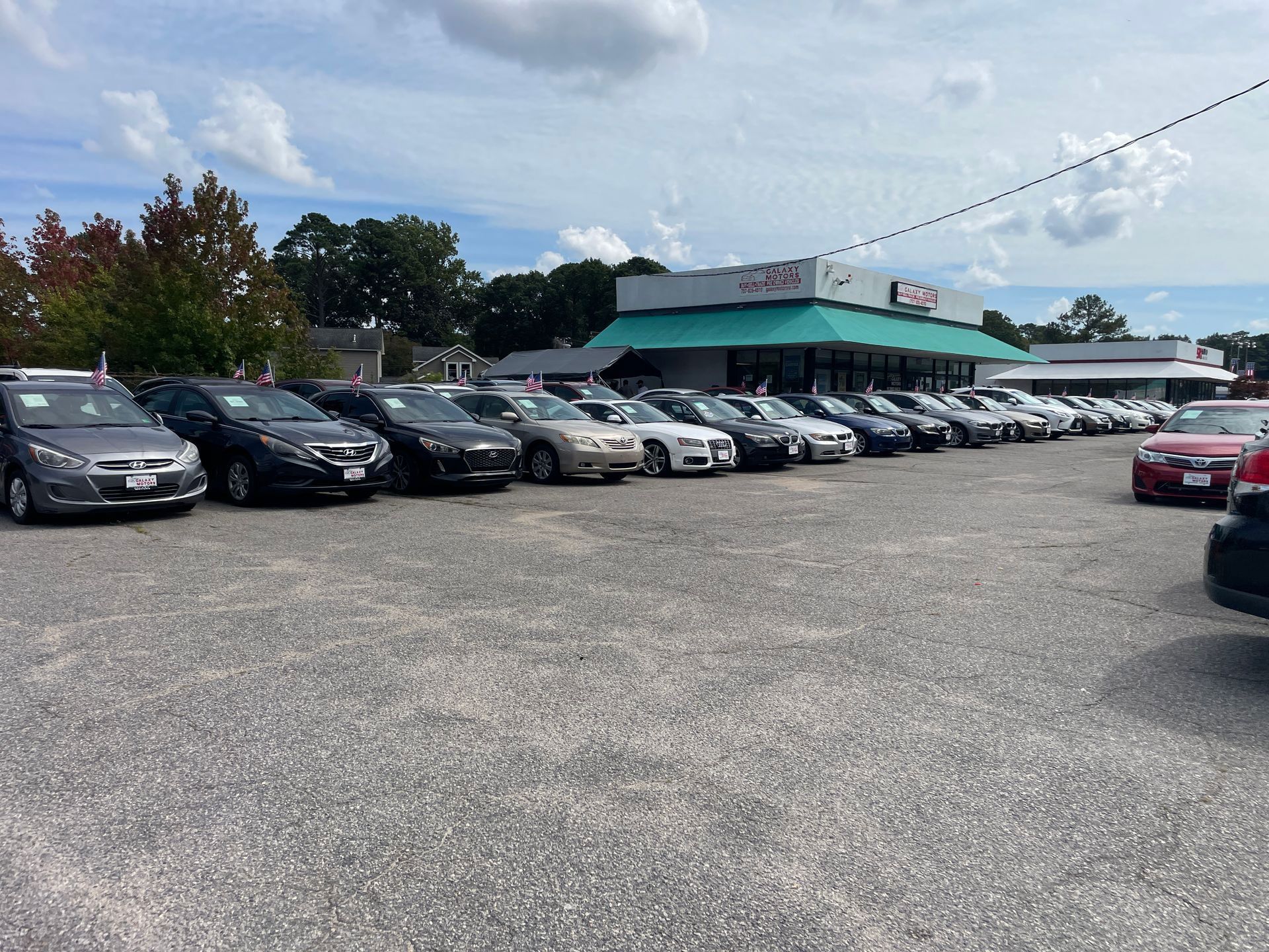 Cars parked in rows at a car dealership under a cloudy sky. Building in the background with a green awning.