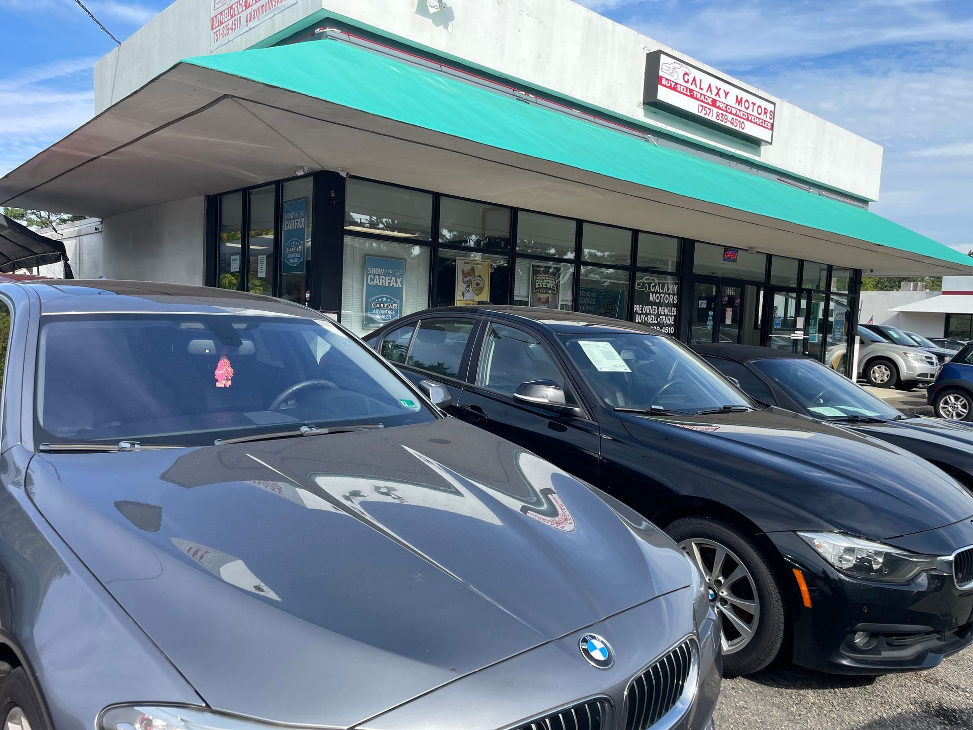 Cars parked in front of a car dealership with a white building and turquoise awning.