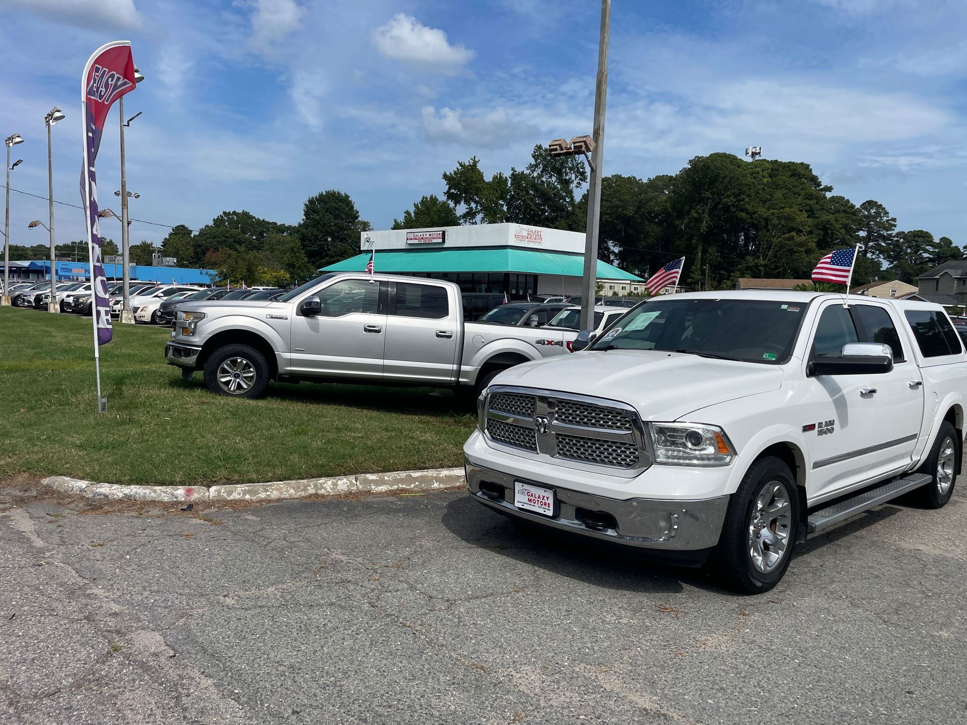 Trucks parked on a gravel lot, a white Ram in the foreground, a silver Ford F-150 behind it, dealership in background.