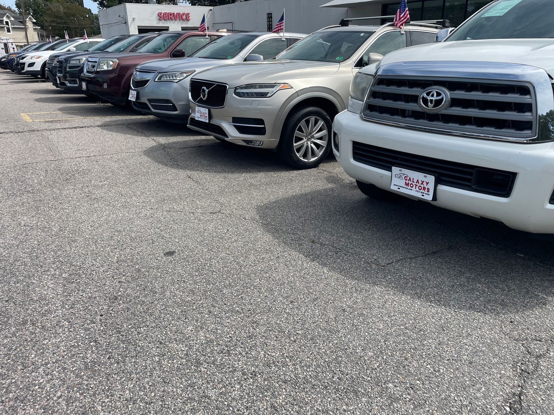 Cars parked in a dealership lot, showing a variety of models in neutral colors under a sunny sky.