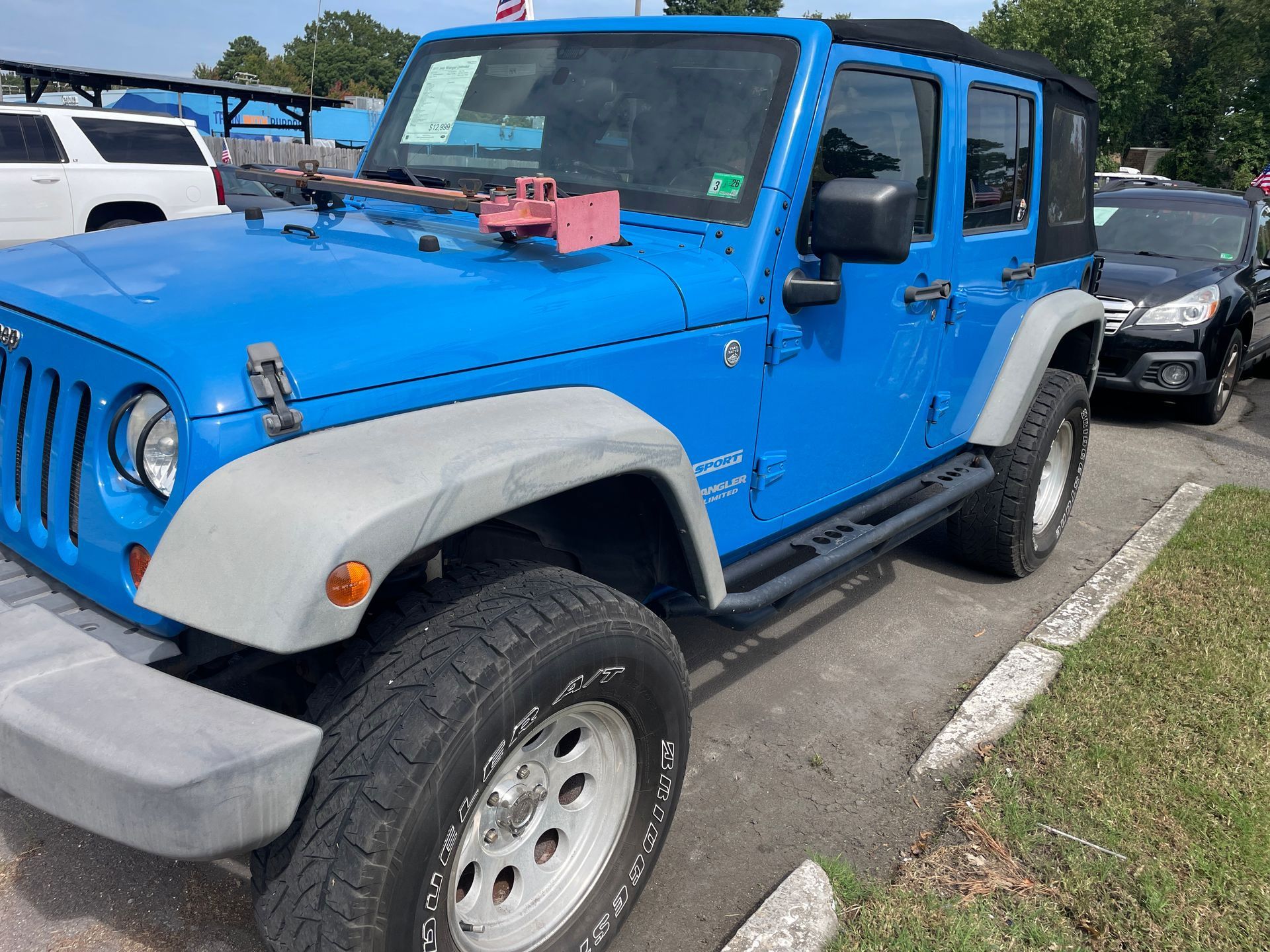 Blue Jeep Wrangler parked in a car lot. It has a black soft top, gray fenders, and a roof rack.