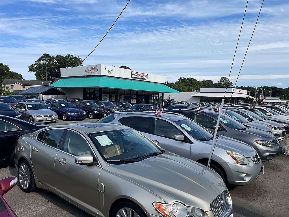 Cars parked at a used car dealership on a sunny day; building with a green awning in the background.
