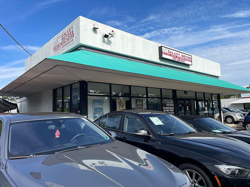 Cars parked in front of a used car dealership with a white building and green awning under a blue sky.