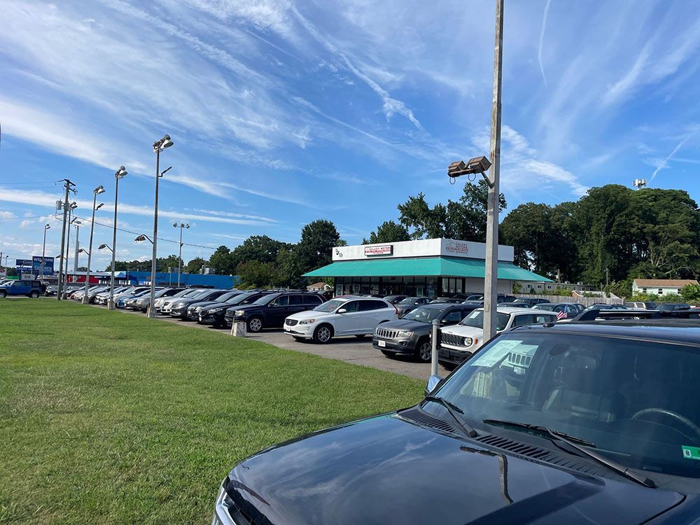 Cars parked at a dealership under a blue sky with a building and street lights.