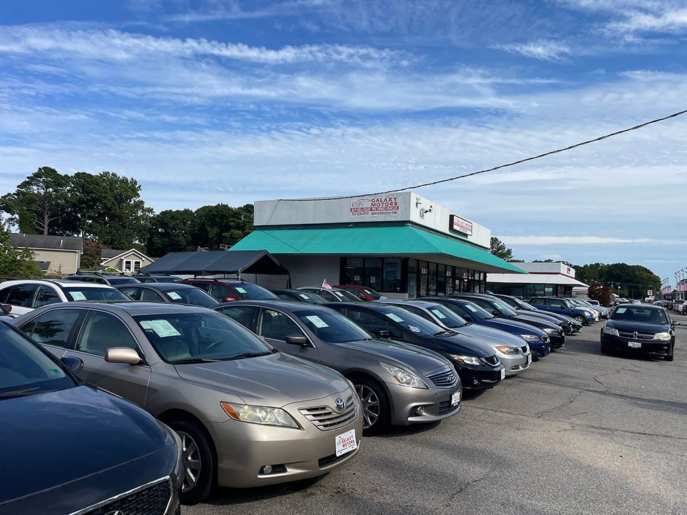 Cars parked at a dealership under a partly cloudy blue sky. The dealership has a green awning.