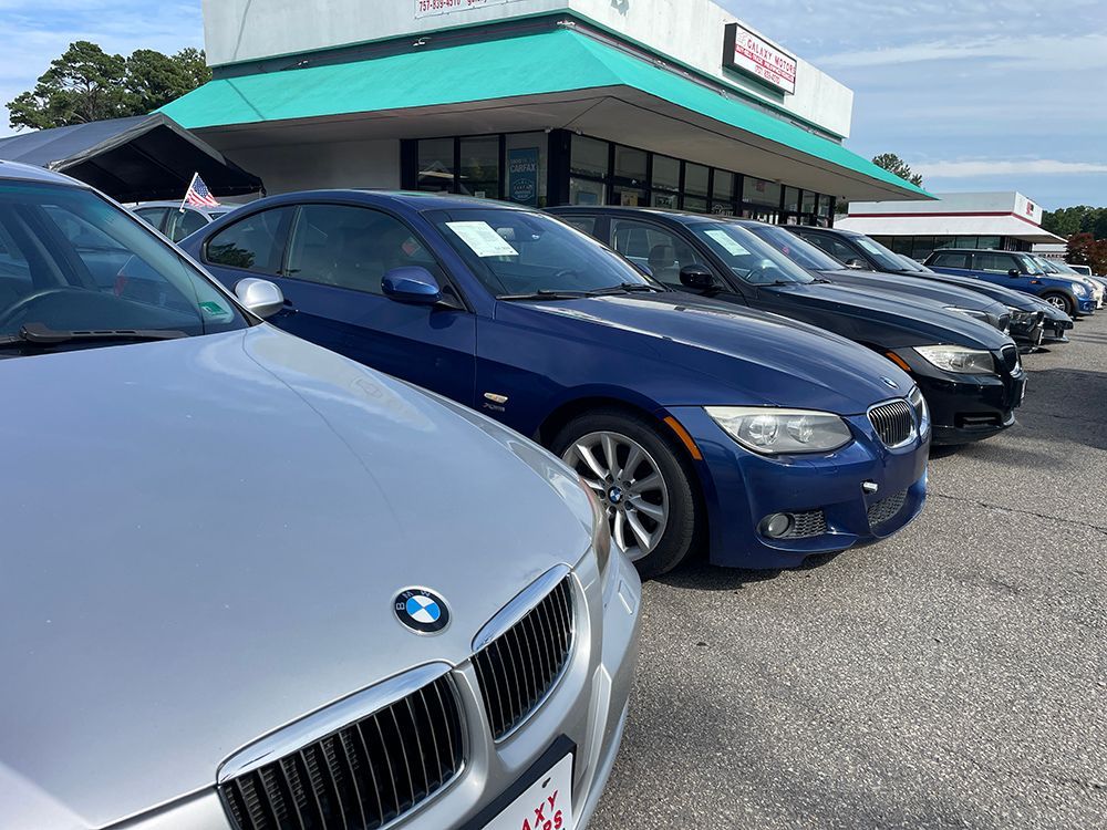 Cars parked in a row in front of a car dealership, including a silver and blue BMW, on a sunny day.