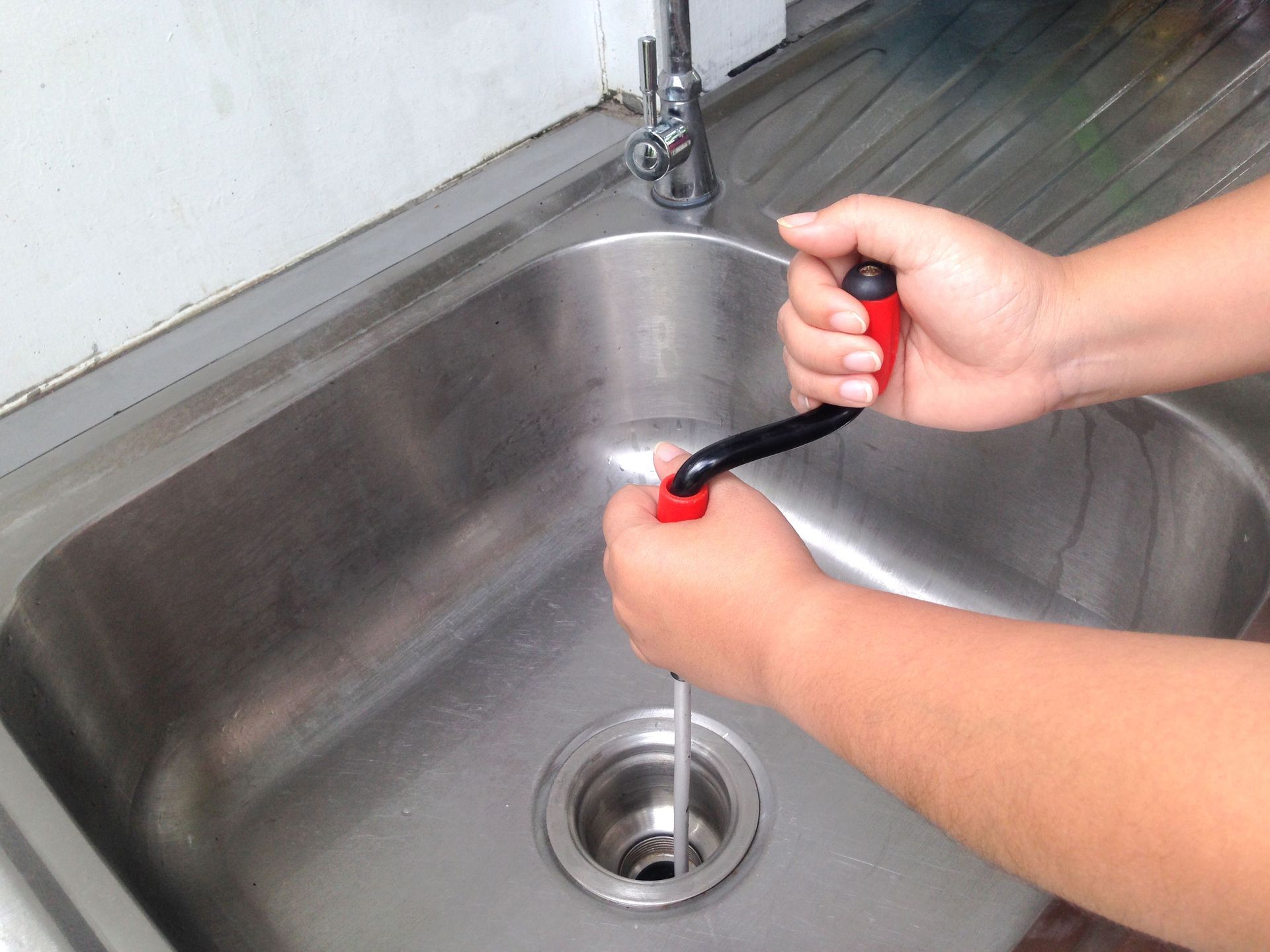 Hands using a red-and-black drain auger to unclog a stainless steel sink.