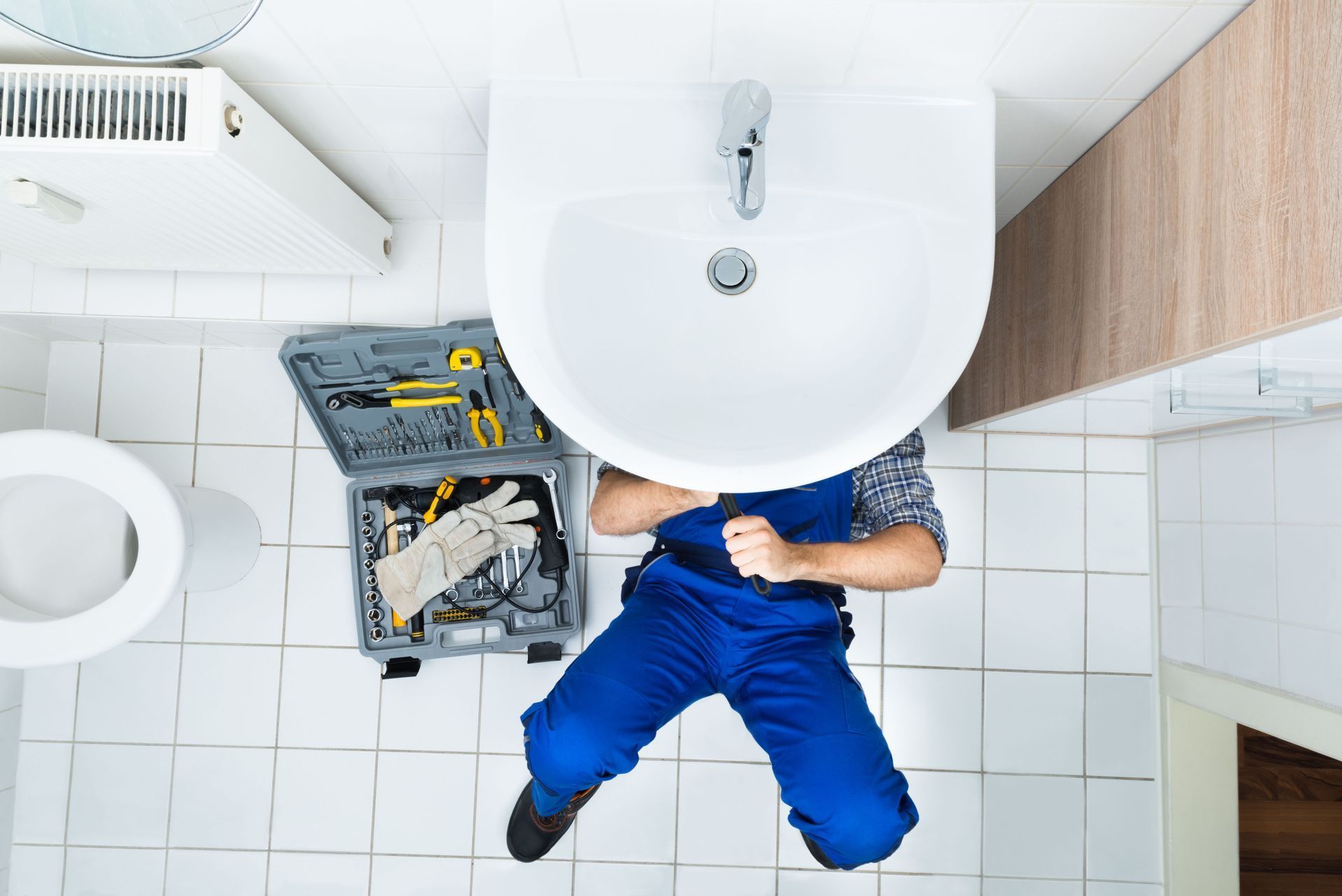 Person in blue clothes lying on white tile floor beside open tool case under a bathroom sink