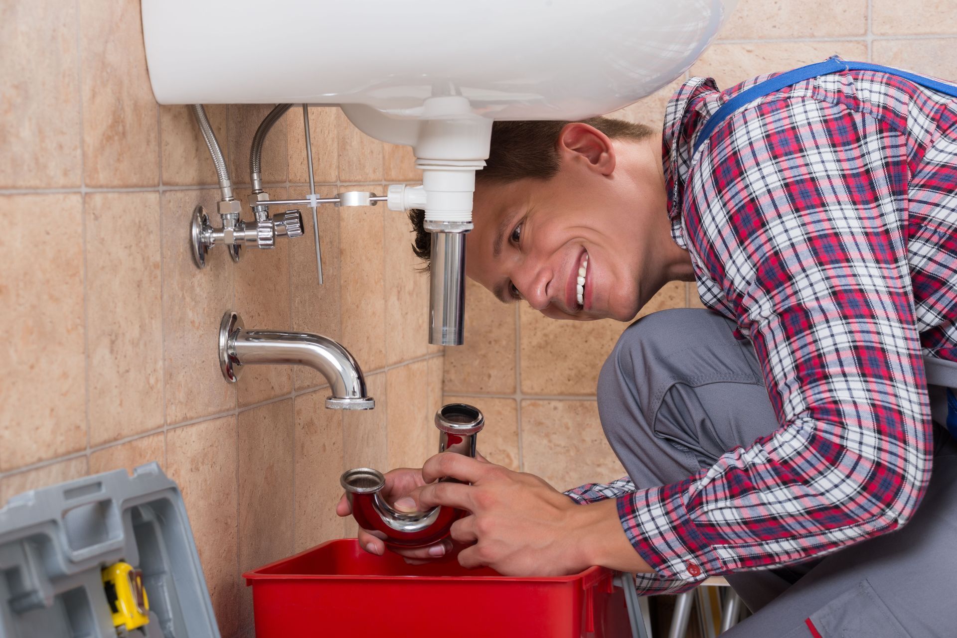 Plumber fixing a sink pipe under a bathroom sink, using a red toolbox and smiling at the camera