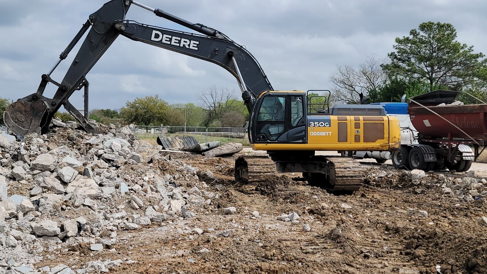 A yellow John Deere excavator works on a pile of rock and rubble at a construction site next to a dump truck.