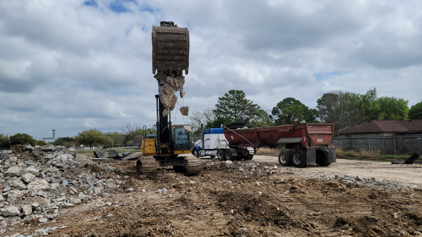 An excavator lifts a bucket of debris at a construction site next to a dump truck under a cloudy sky.