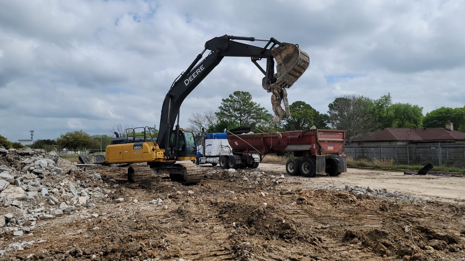 A yellow and black excavator loads rubble from a construction site into a red dump truck under a cloudy sky.
