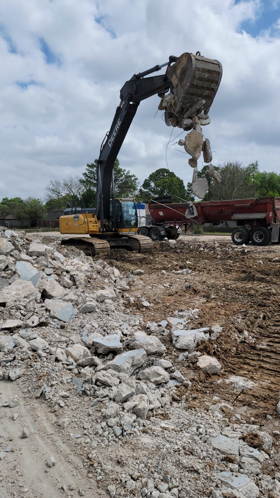A yellow and black excavator lifts concrete debris over a work site with a dump truck nearby under a cloudy sky.