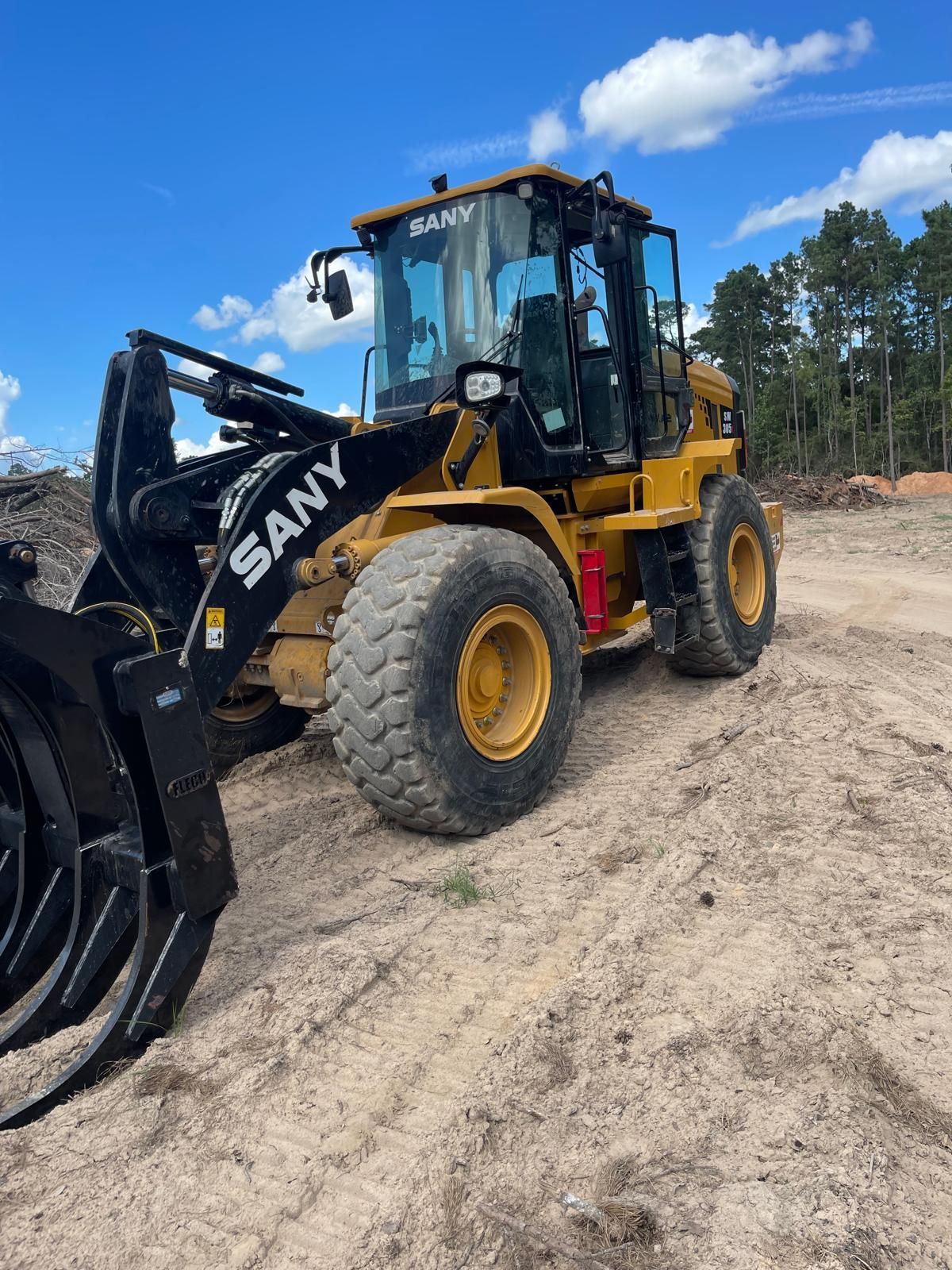 A yellow Sany wheel loader with a log grapple attachment sits on a dirt site under a blue sky.