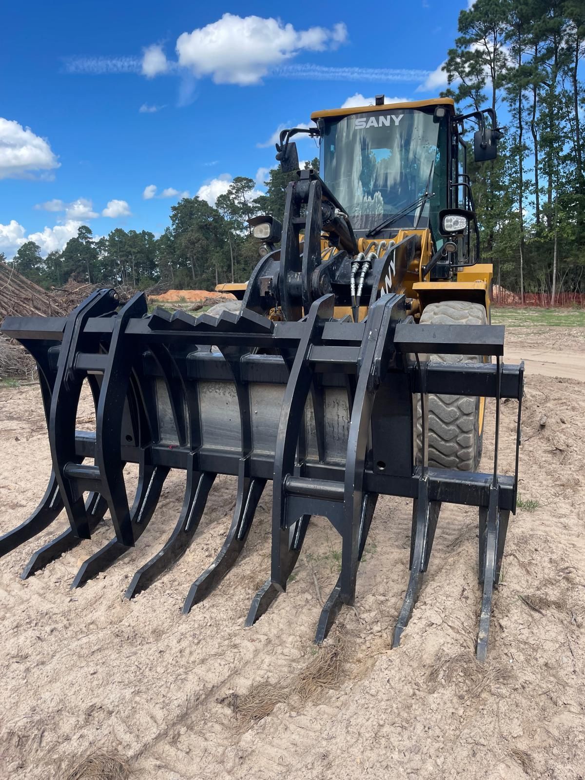 A yellow SANY wheel loader with a large, black metal log grapple attachment parked on a dirt lot under a blue sky.