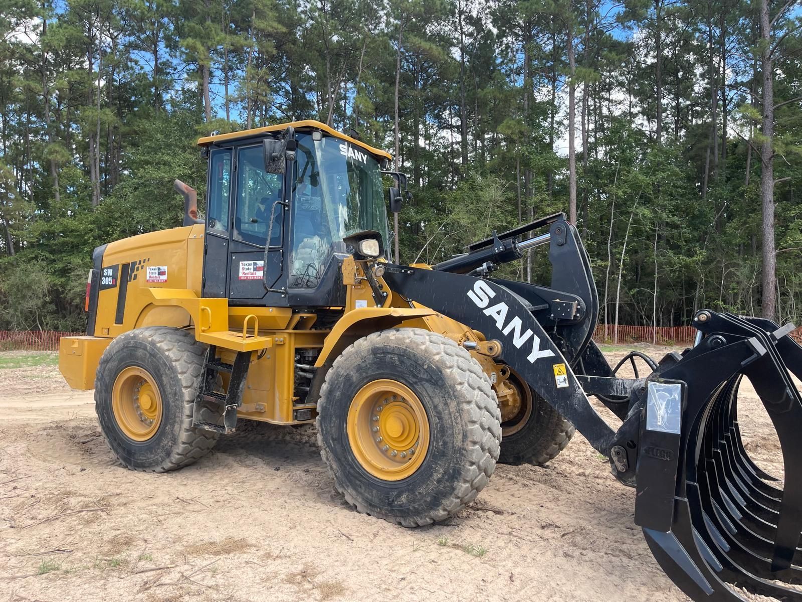 A yellow SANY wheel loader with a large black grapple attachment parked on a dirt lot with a treeline in the background.