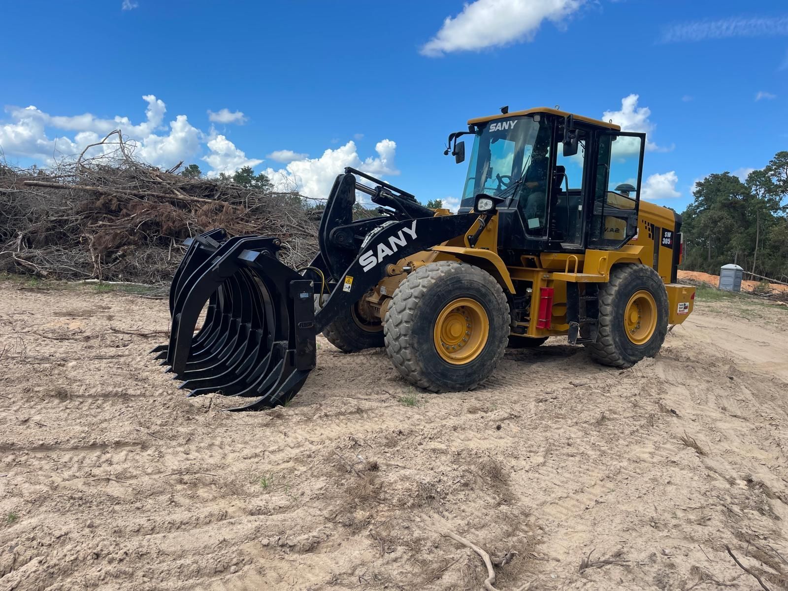 A yellow Sany wheel loader with a large black grapple attachment sits on a dirt construction site under a blue sky.