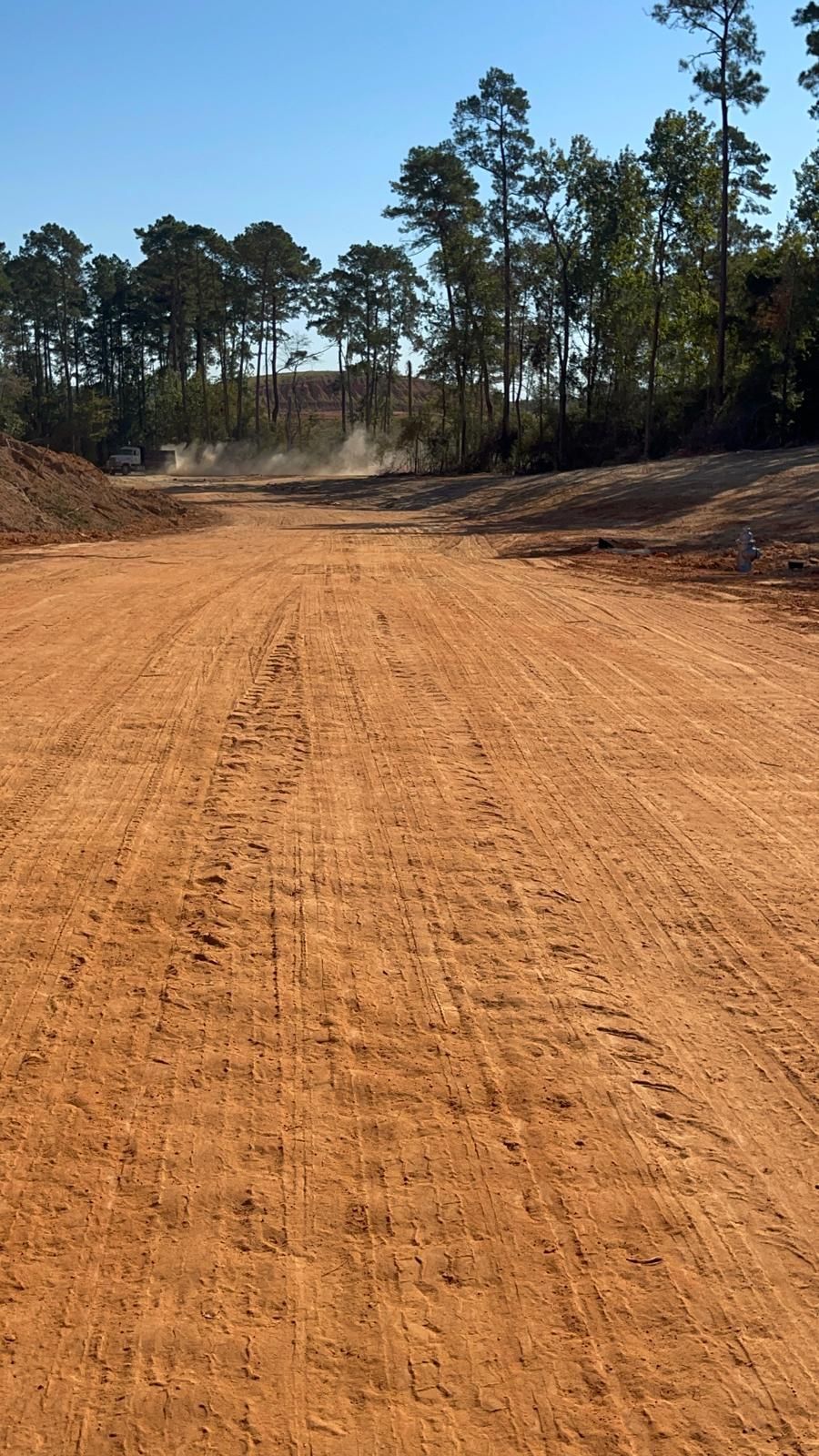 A dirt path winds through a construction site, flanked by mounds of earth and trees under a clear blue sky.
