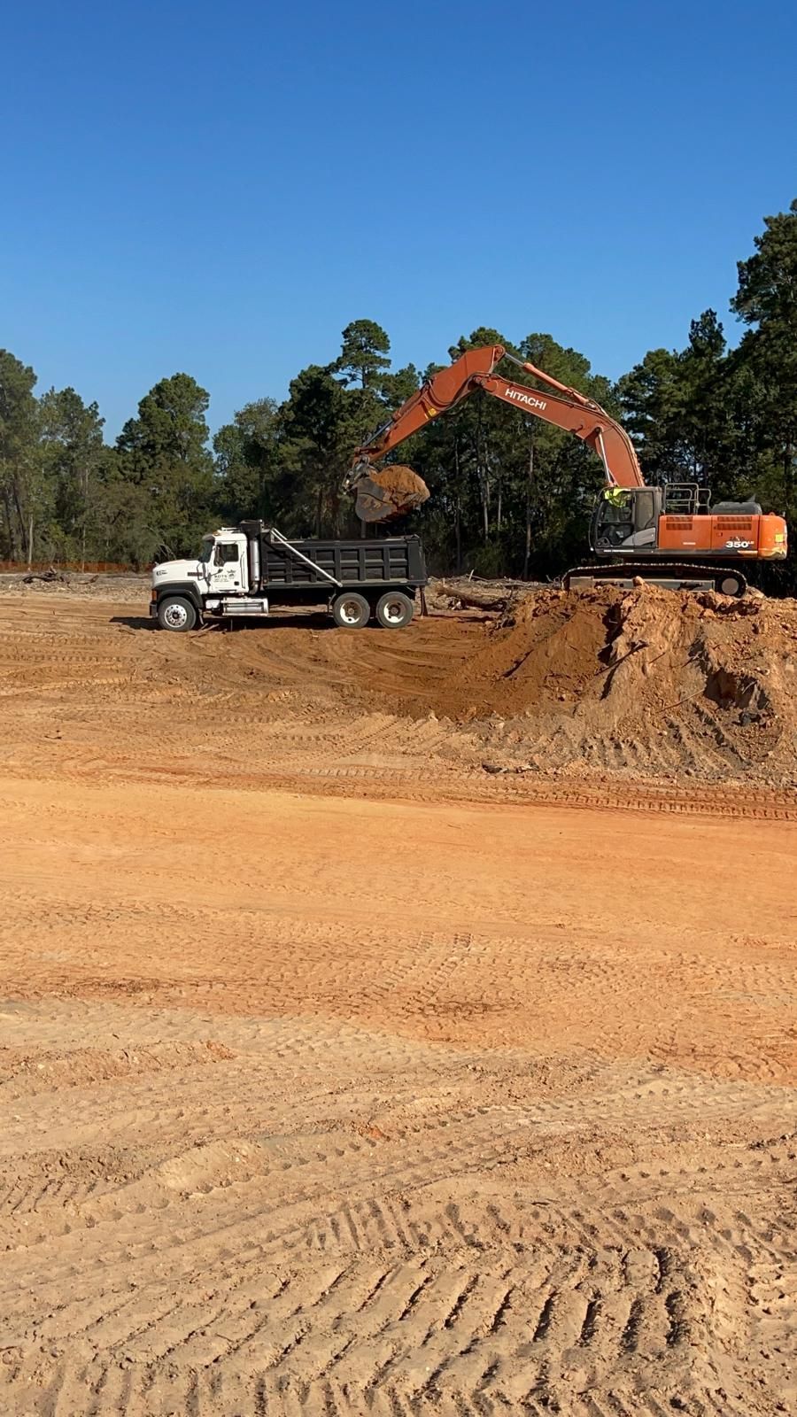 An orange excavator loads dirt into a white dump truck at a construction site with trees in the background under a blue sky.