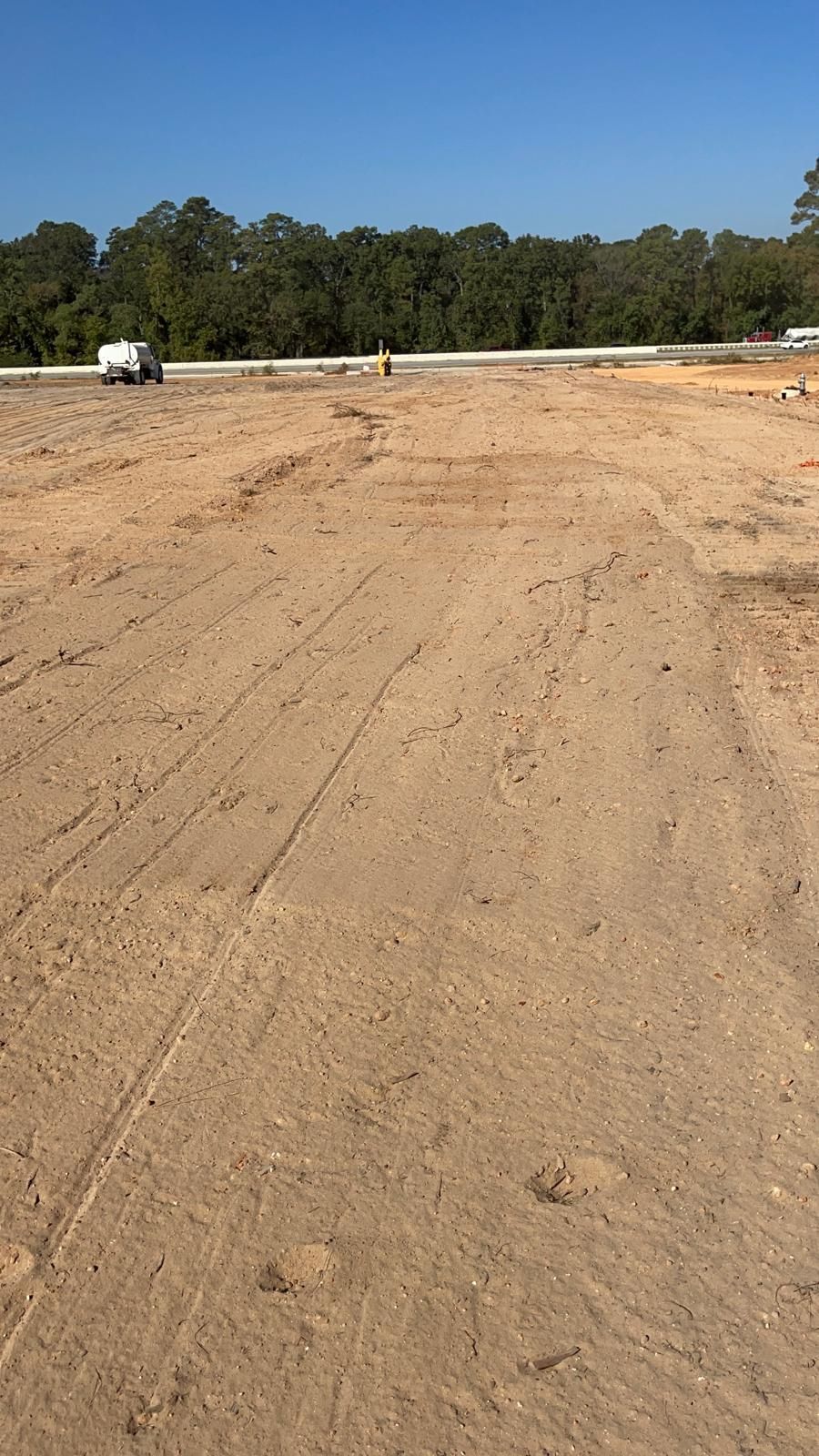 A large, cleared construction site with dirt tracks, a white vehicle in the distance, and a forest line under a blue sky.