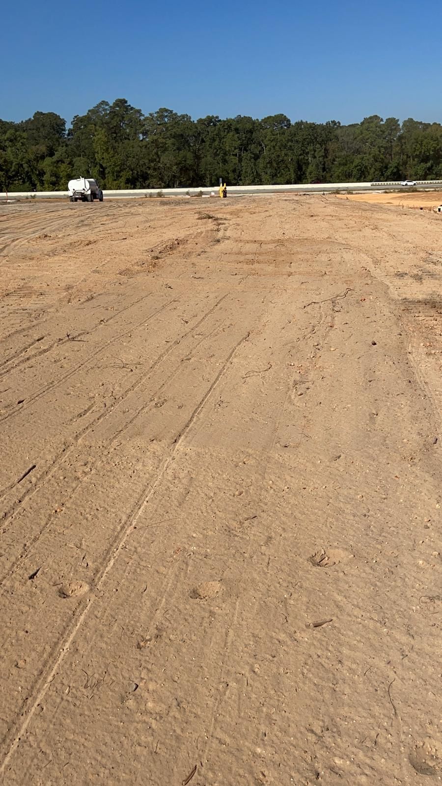 A wide, dirt-covered construction site with tire tracks leading toward a distant road, white truck, and treeline.
