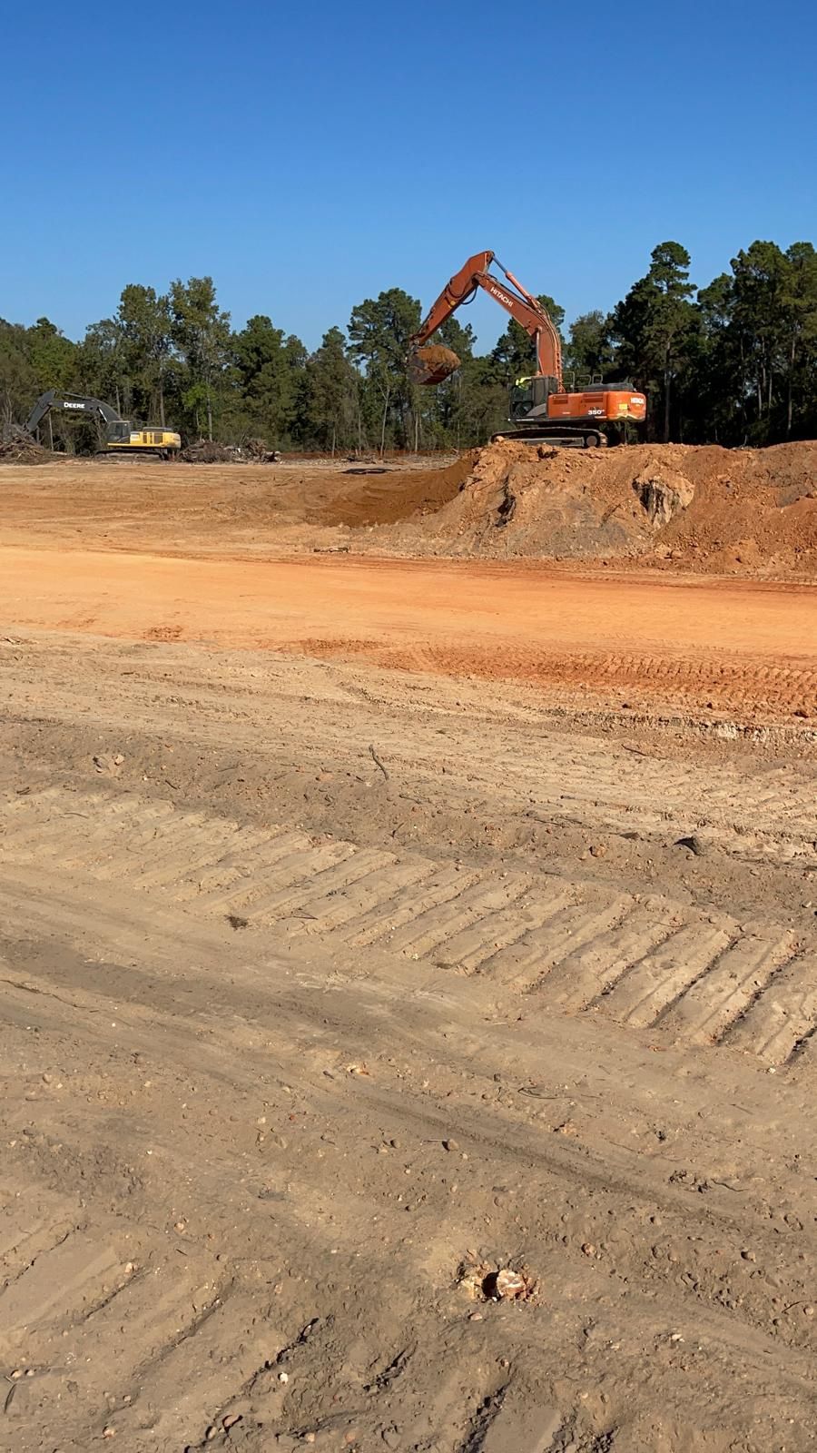 An orange excavator sits on a dirt mound at a construction site with another piece of equipment nearby under a blue sky.