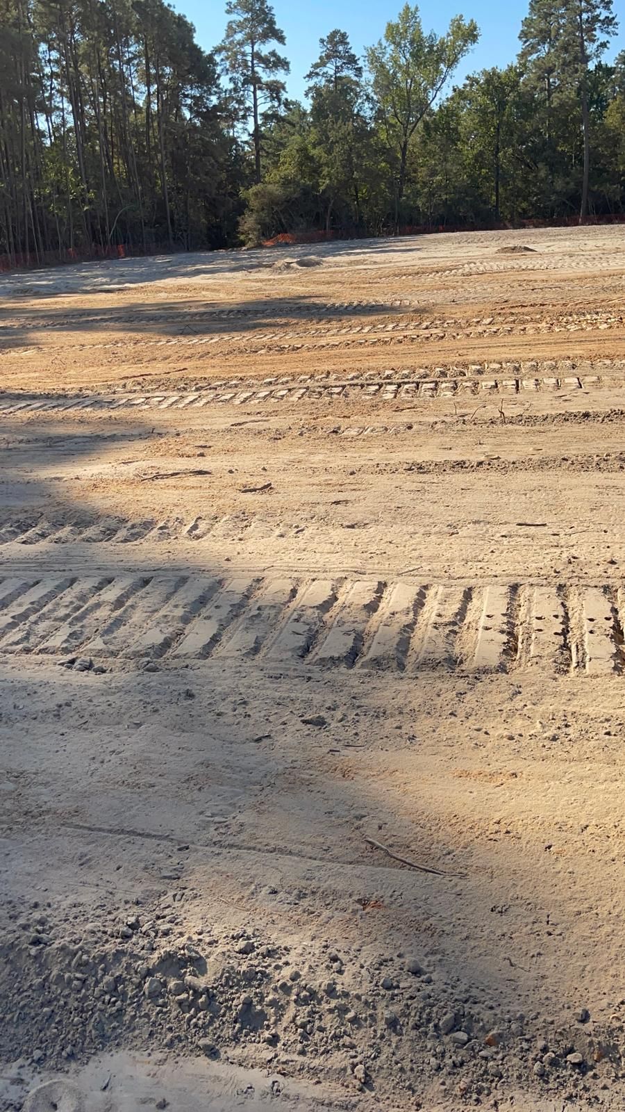 A wide view of a graded dirt lot with distinct heavy equipment tread marks across the sandy, dry ground near tree lines.