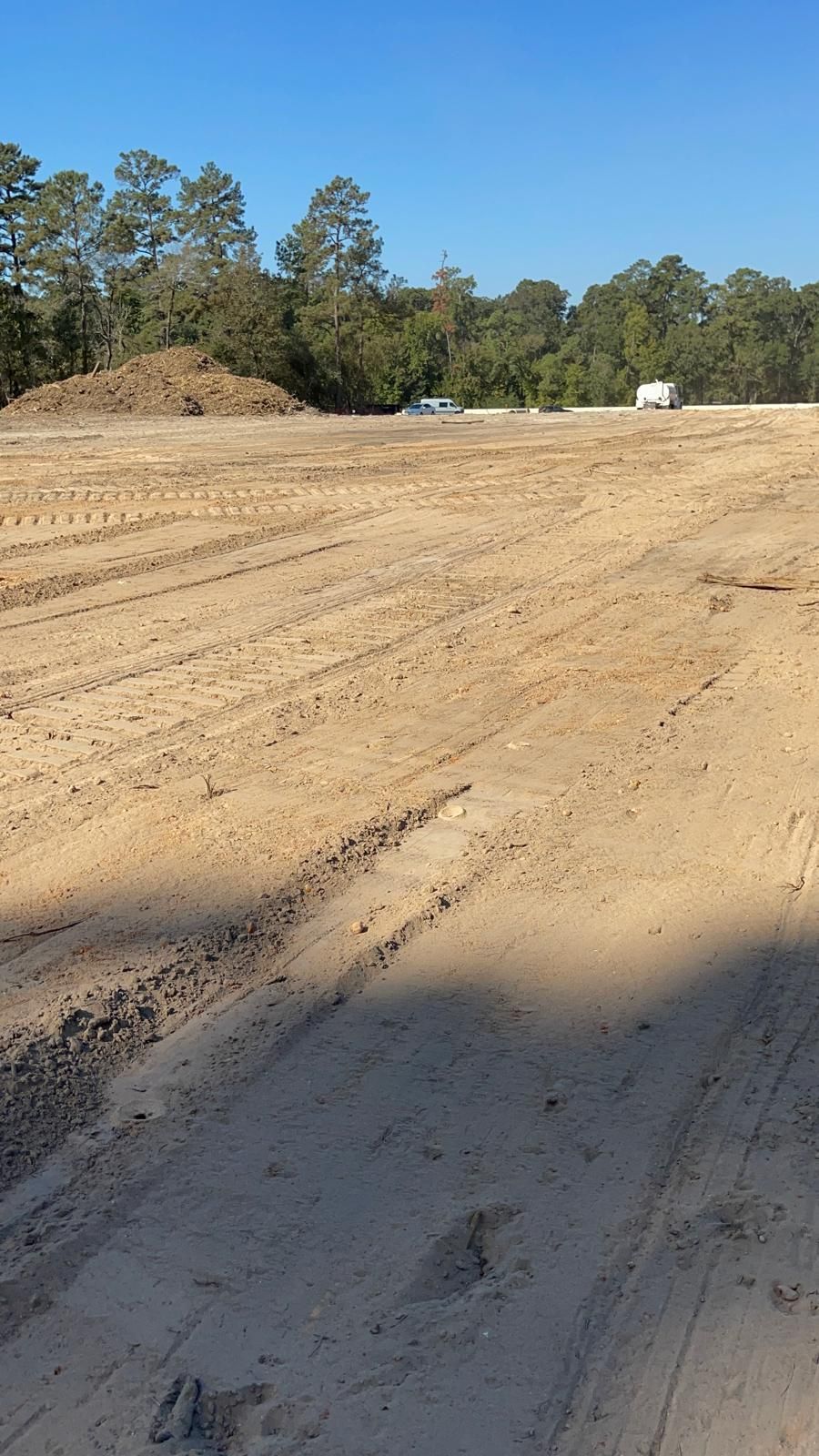 A wide, sunny construction site with cleared sandy dirt, tire tracks, a large dirt mound, and distant trees.