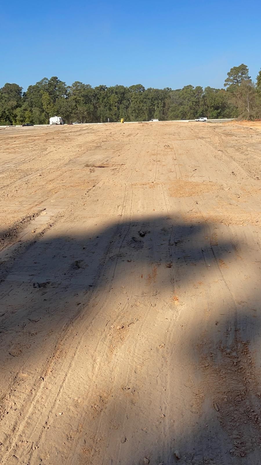 A flat, cleared dirt construction site with tire tracks, under a clear blue sky, facing a distant treeline.