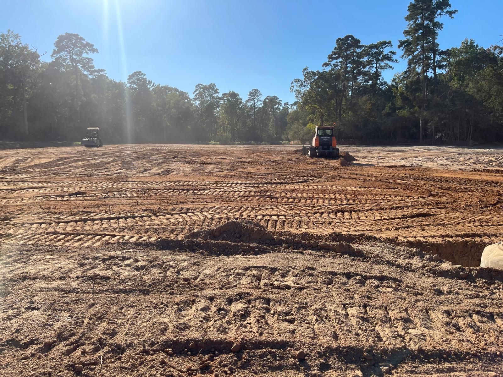 Two excavators clear land on a sunny, dirt-covered construction site with a forest in the background.