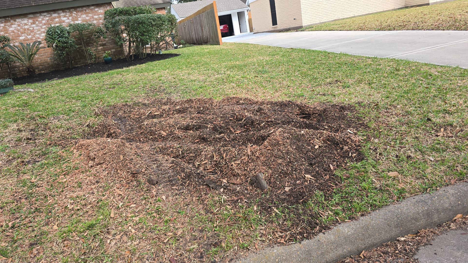 A patch of dark mulch surrounded by green grass in a suburban front yard.