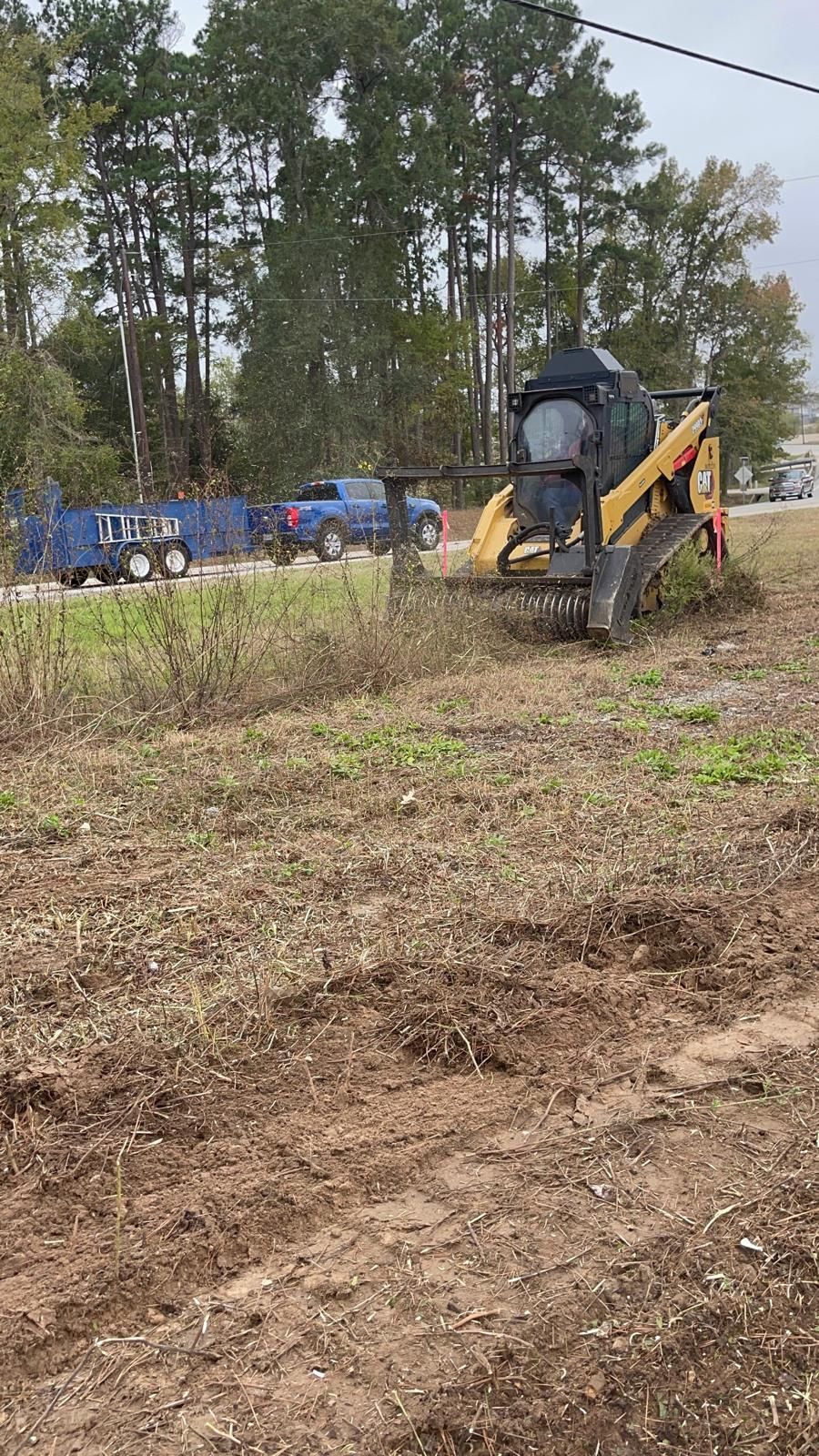 A yellow skid steer mower clears brush and tall weeds in a grassy field with a blue truck parked in the background.