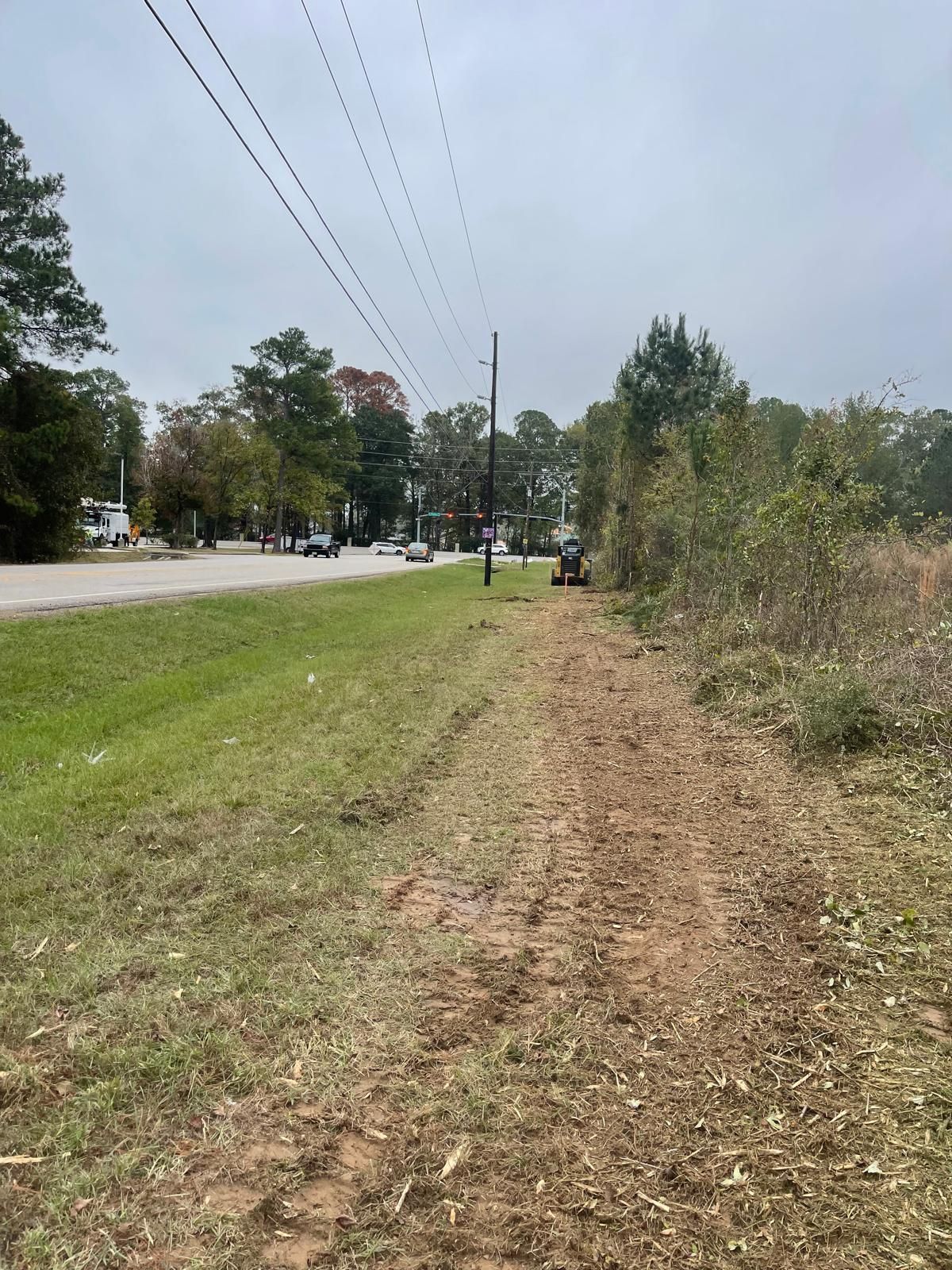 A tractor clearing vegetation along a roadside under power lines on an overcast day.