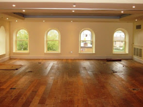 Empty hardwood-floored room with arched windows and beige walls, lit by recessed ceiling lights