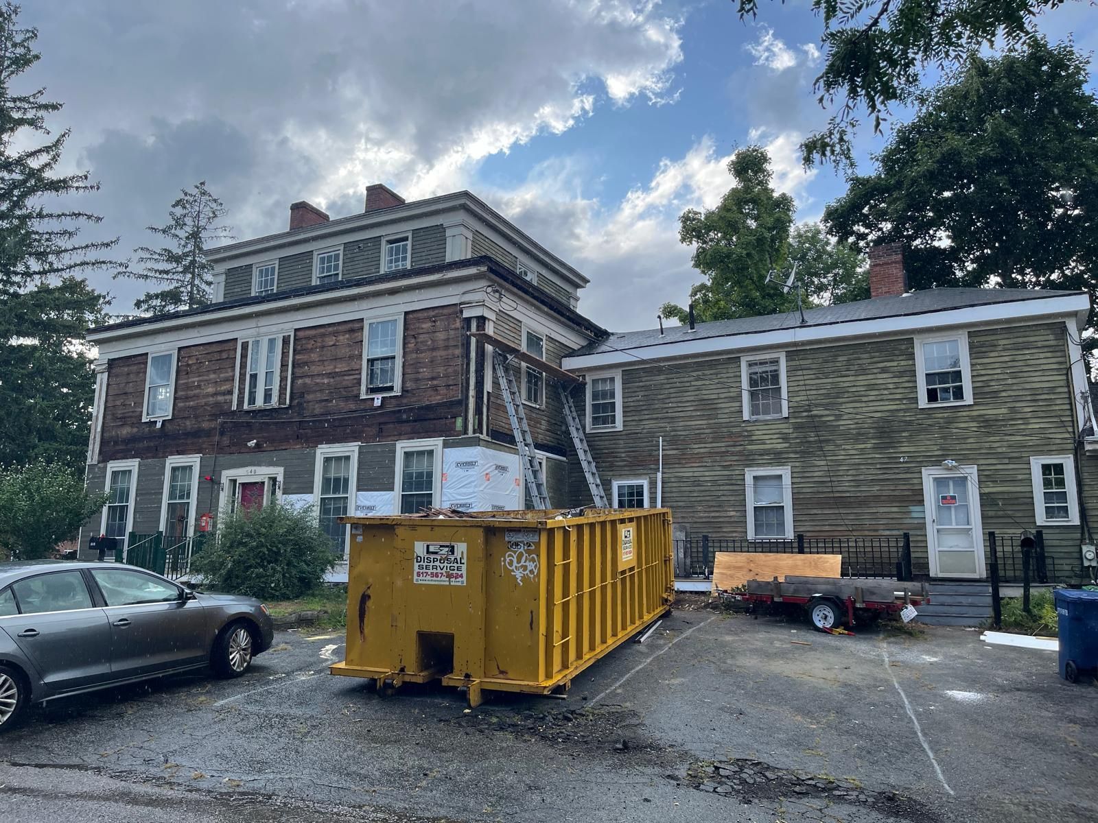 Two-story houses and a yellow construction dumpster in a gravel driveway under cloudy skies