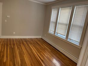 Empty room with polished hardwood floors, beige walls, and three large windows with blinds.