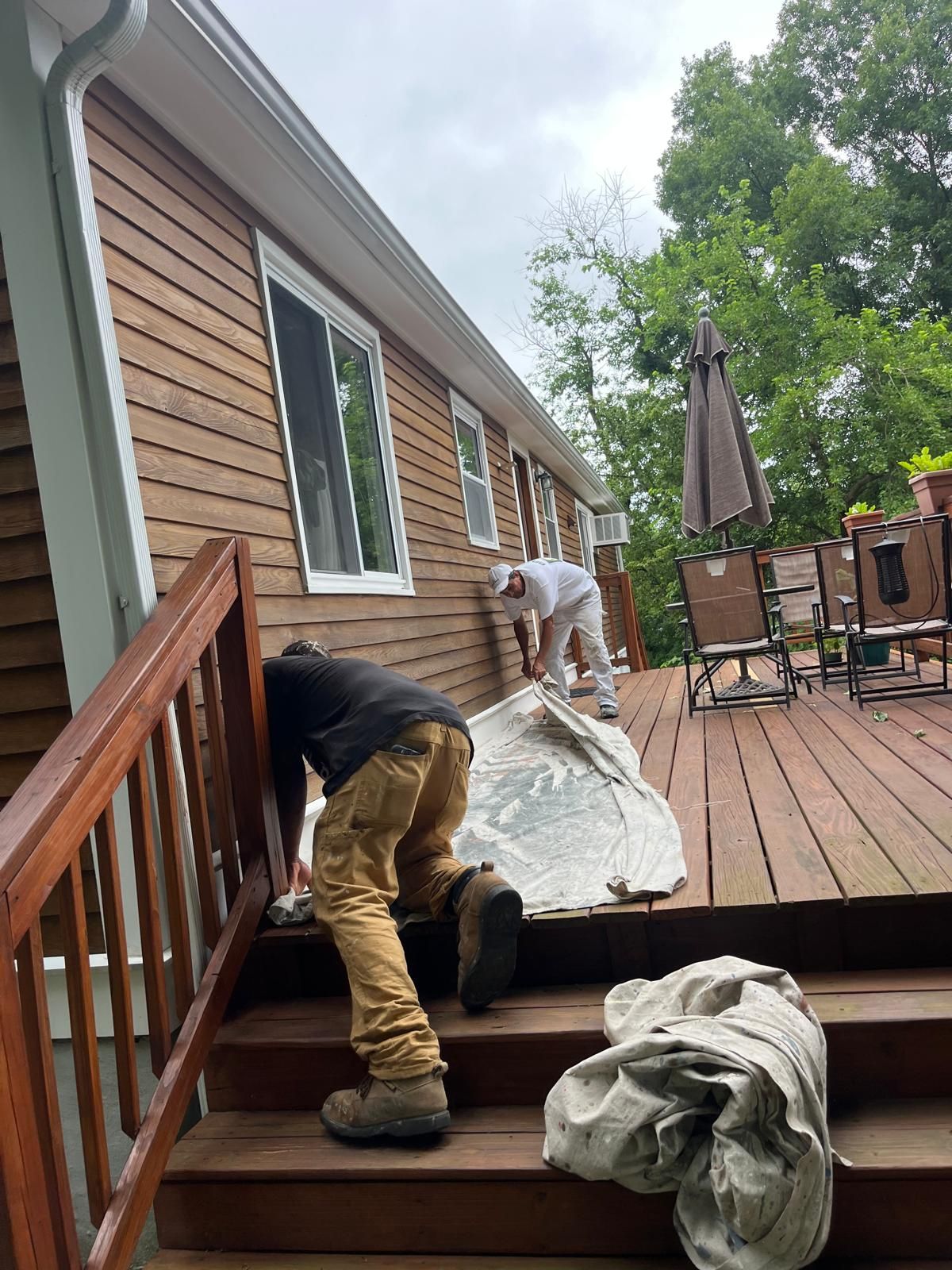 Two workers moving a large wrapped item across a wooden deck beside a house
