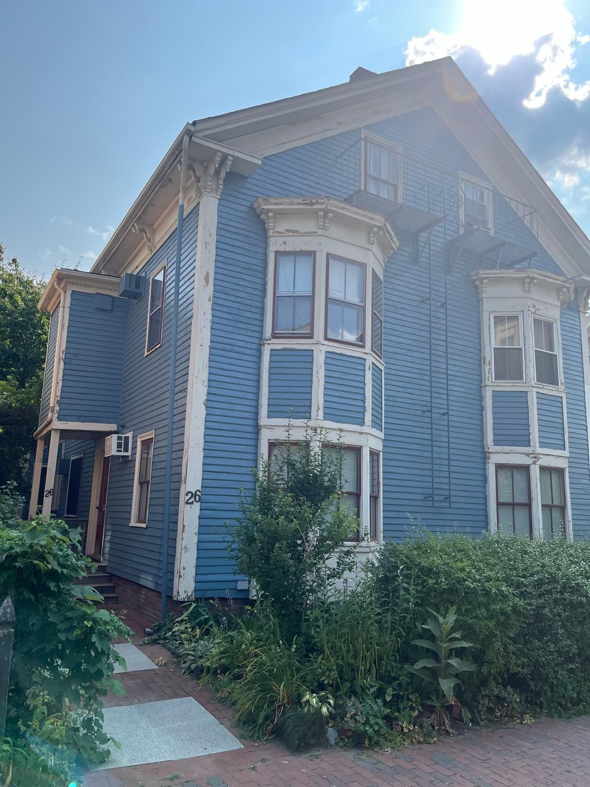 Blue Victorian house with bay windows and overgrown greenery beside a sidewalk under a bright sky