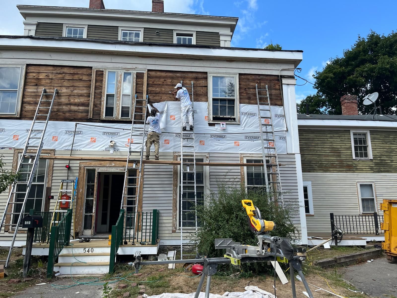 Workers on scaffolding repairing the white exterior of a weathered house with blue trim