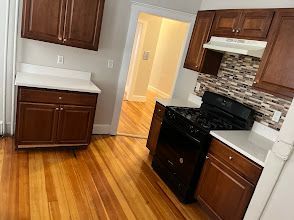 Empty kitchen with dark wood cabinets, black stove, white counters, and hardwood floors near a doorway.